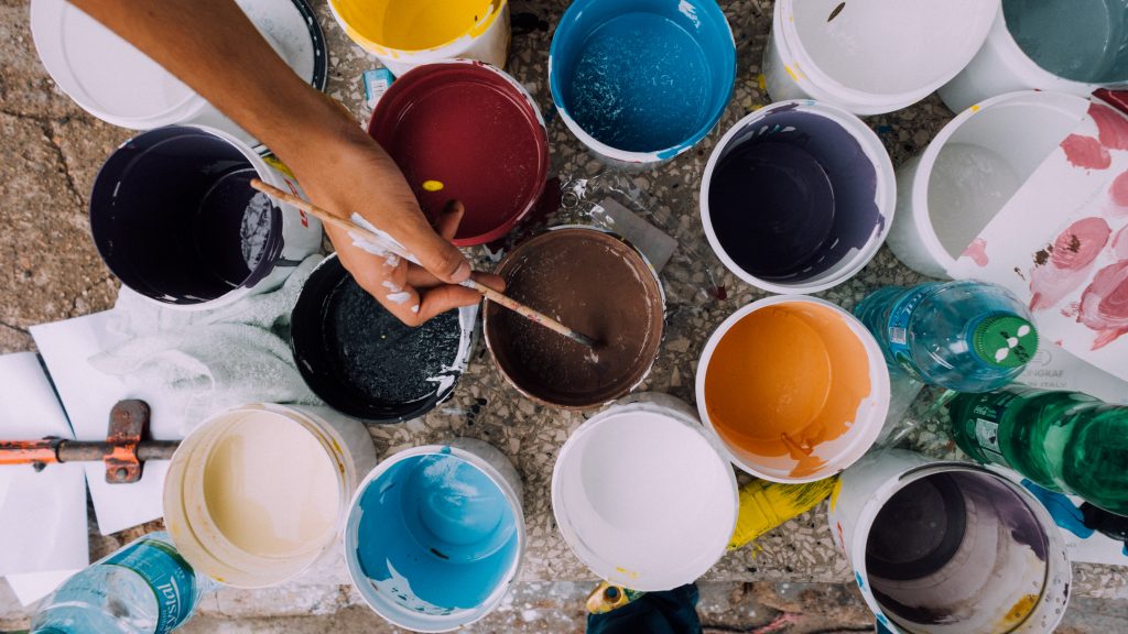 A woman dips a paintbrush into a can of paint. 